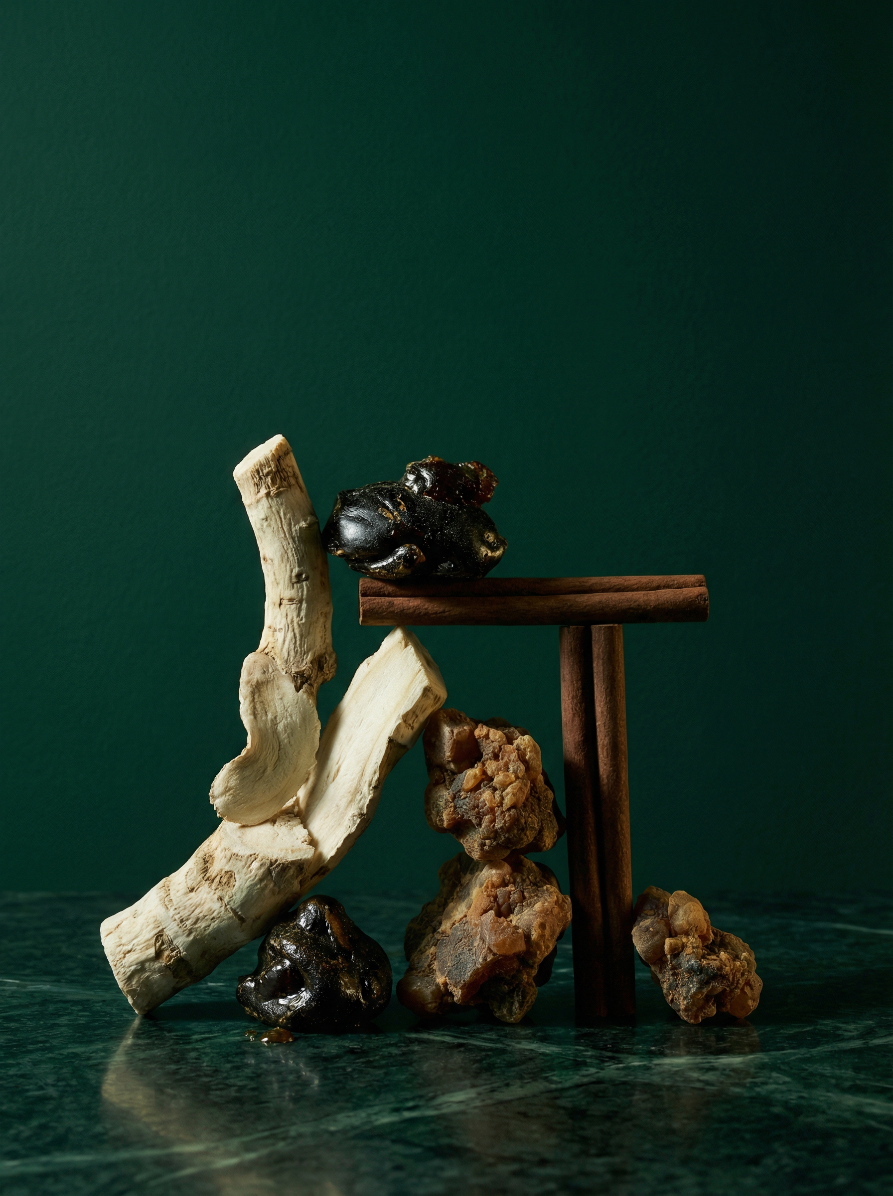 Heart note ingredients of Code of Wealth perfume — orris root, labdanum resin, myrrh and cinnamon sticks on dark emerald surface