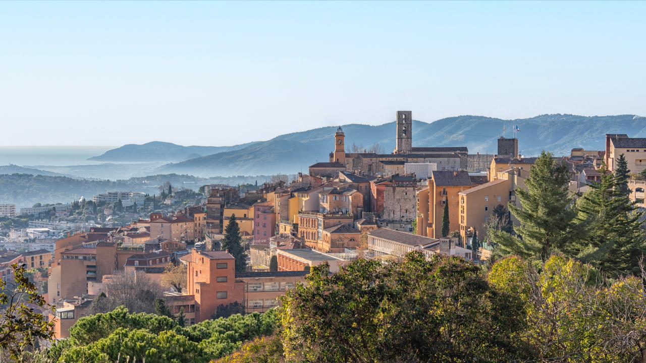 Panoramic view of Grasse, France — the world capital of perfumery — with historic terracotta buildings, church tower and misty mountains in the background